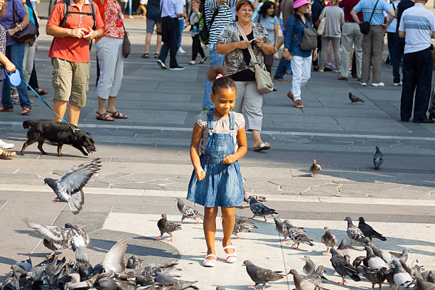 Milan, Italy - September, 1st 2013: Scene on square Piazza Duomo with little black girl standing between pidgeons. In background are many tourists doing sight seeing between Galleria Vittorio Emanuele II and cathedral Maria Nascente.