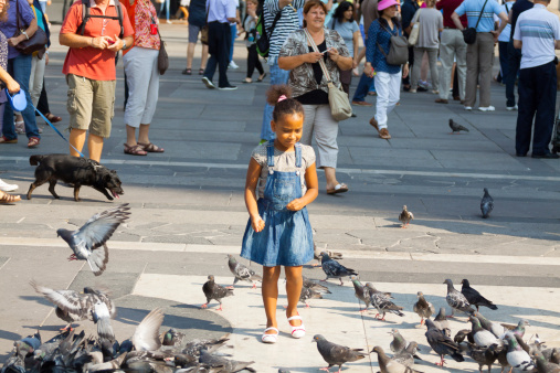 Milan, Italy - September, 1st 2013: Scene on square Piazza Duomo with little black girl standing between pidgeons. In background are many tourists doing sight seeing between Galleria Vittorio Emanuele II and cathedral Maria Nascente.