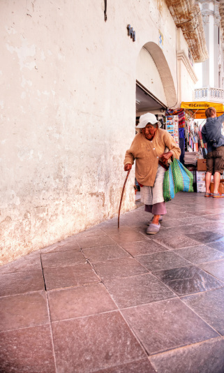 Arequipa, Peru - May 25, 2013: Senior Peruvian woman with walking stick and shopping bag  on Arequipa sidewalk. People are pictured in the background