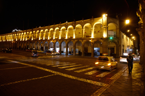 Arequipa, Peru - May 26, 2013: Peruvian arcade surrounding town square at night. People are pictured walking by and cars and taxis are passing and parked at the kerb