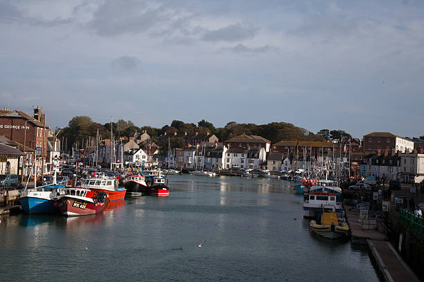 Harbour at Weymouth in Dorset; Weymouth, England - October 25, 2013: Harbour at Weymouth in Dorset;  Small Yachts and fishing boats are moored. Water front properties line the harbour. People can be seen in the image.. Blue sky and blue water with reflections weymouth dorset stock pictures, royalty-free photos & images