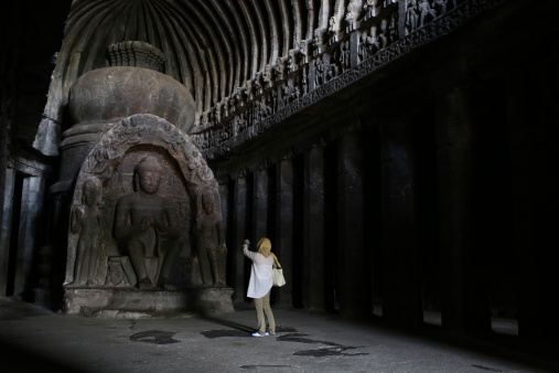 Ellora India - November 15, 2012: Foreign tourist takes a photo of a buddihst statue is carved at the Ellora Caves in Aurangabad district of Maharashtra.  Ellora is an archaeological site, 29 km (18 mi) North-West of the city of Aurangabad in the Indian state of Maharashtra built by the Rashtrakuta dynasty.  Well known for its monumental caves, Ellora is a World Heritage Site.  Ellora represents the epitome of Indian rock-cut architecture. The 34 