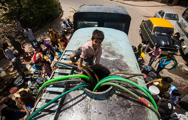 New Delhi, India - May 21, 2013: An Indian boy sits atop a government water supply truck as residents fill water containers. During the hot summers residents struggle with water shortages. In many poor parts of New Delhi there is limited running water, and people need water tank deliveries to beat the heat.