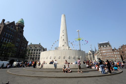 Amsterdam, the Netherlands - April 22, 2011: The National Monument at Dam Square in Amsterdam. A carnival with a ferris wheel and a chain swing ride at the background.  am Square lies in the historical center of Amsterdam and is one of the city's most well known places. It's a meeting place and a location for many events. The National Monument is a Second World War monument. In the picture, people are sitting on the monument, relaxing in the sun.