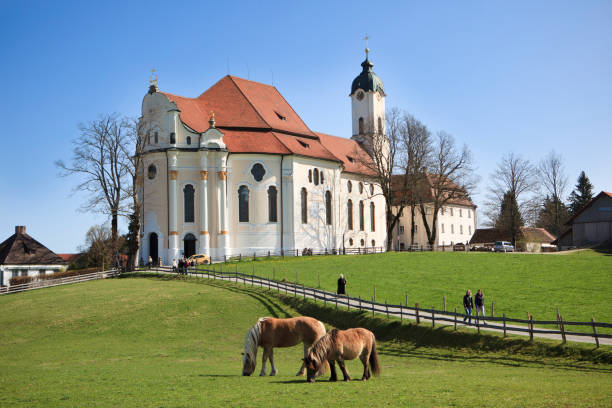 Steingaden, Germany - April 10, 2011: Worshippers leaving the Pilgrimage Church of Wies (Wieskirche) after Sunday mass. The church of Wies is an oval Rococo Church near Steingaden at the foothills of the Alps. It was designed by Dominikus Zimmermann in the 1740s and added to the UNESCO World Heritage List in 1983. Two ponies grazing on the meadow in the foreground.