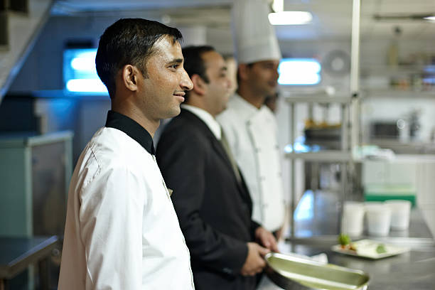 Delhi, India - November 12, 2010: Two Indian hotel chefs preparing food in a busy Delhi hotel kitchen. Four otrher chefs and maitre d'hotel can be seen standing waiting in the background