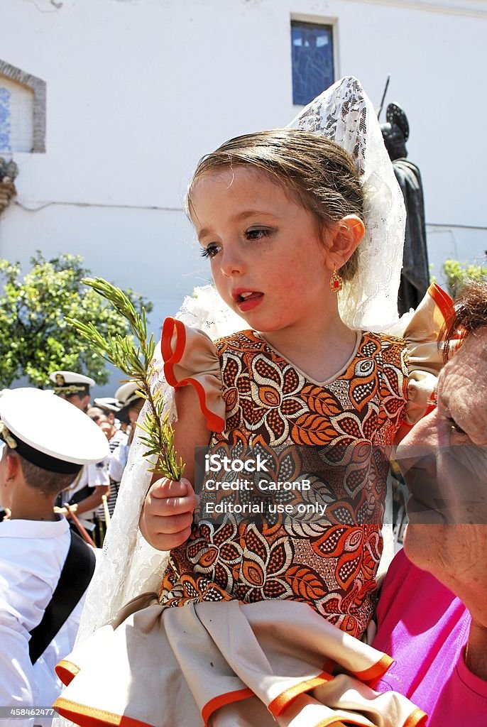 Small girl holding sprig of rosemary for luck, Marbella, Spain. Marbella, Spain - June 11, 2008: Girl in traditional dress being held in her grandmother's arms carrying a sprig of rosemary in the Plaza de la Iglesia, Romeria San Bernabe, Religious Festival, Marbella, Spain. Adult Stock Photo