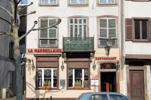 Strasbourg, France - February 22, 2013: The Restaurant La Marseillaise at the Place Broglie in the city of Strasbourg, Alsace, France. Picturesque, old facade. Menues, billboards and advertising at the entrance and the window. One man is visible through the window. Roof of a parking car in the foreground. Sunny winter day.