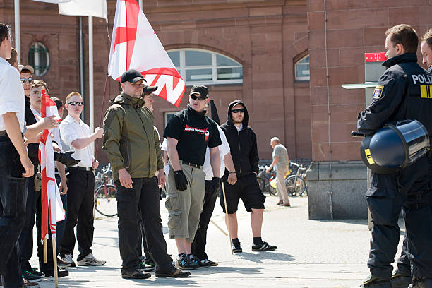 Young german Neonazis against riot police Wiesbaden, Germany - May 28, 2011: A group of young german Neonazis - prepared to use violence - is being obeserved by members of german riot police in the center of Wiesbaden during a Neonazi-march of JN (Junge Nationaldemokraten). Junge Nationaldemokraten is the youth organisation of NPD (Nationaldemokratische Partei Deutschlands). Founded in 1964 the NPD is a German nationalist party, its agitation is racist, antisemitic, revisionist. Because of its activities against the constitutional order the party is under observation of the Verfassungsschutz (German Federal Intelligence agency). In the background a non-involved pedestrian. national democratic party of germany stock pictures, royalty-free photos & images