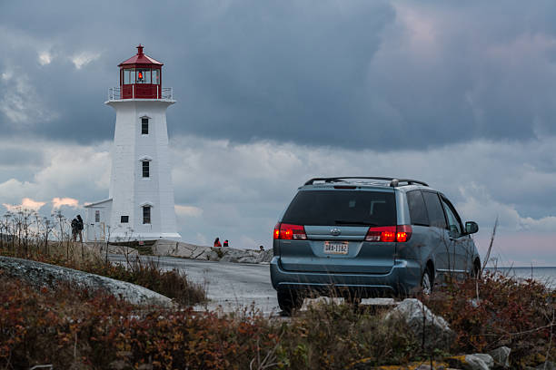 Peggy's Cove, Canada - October 27, 2013: An american tourist pauses at the end of Peggy's Point Road soaking in the dramatic views of Peggy's Cove Lighthouse under turblent skies in late evening.  A number of other tourists in the background enjoy the view from the base of the lighthouse.