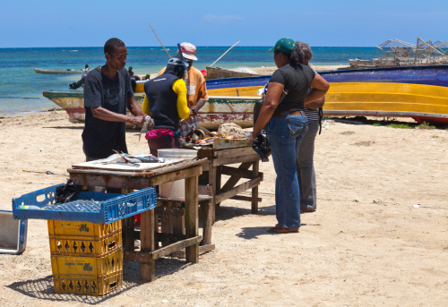 Falmouth, jamaica, August 28, 2013: A group of people standing on a beach, two fishermen one dressed in black and the other wearing a wetsuit, preparing fish to sell to the buyers, two women with jeans and a man with a hat