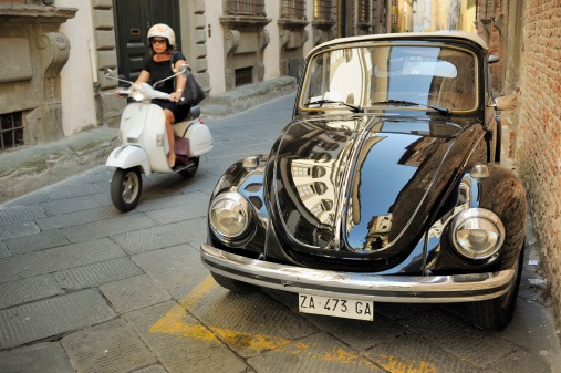 Lucca, Italy - June 27, 2011: A woman on a motor scooter drives past a parked vintage Volkswagen Beetle Convertible car. This special version of the VW classic was manufactured by the Karmann company between 1949 and 1980.