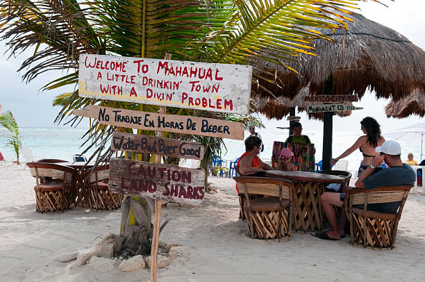 Costa Maya, Mexico - March 24, 2011: Tourists sit at a beachside cafe in this Yucatan resort town, popular with cruise ships. A local man is standing at the table and displaying bracelets for sale. Among the notices on the sign: WELCOME TO MAHAHUAL, A LITTLE DRINKIN TOWN WITH A DIVIN PROBLEM
