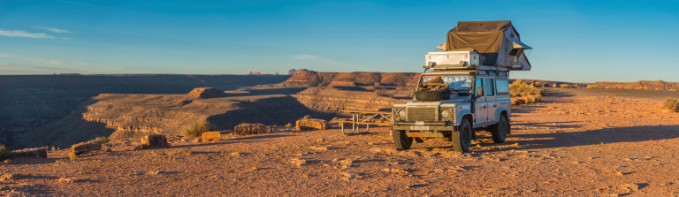 Mexican Hat, Utah, USA - 8th November 2013: Rugged Land Rover Defender with rooftop safari tent and wilderness exploring fixtures camping on the edge of the cliffs at Goosenecks State Park in Monument Valley, Utah, USA. Composite panoramic image created from eleven contemporaneous sequential photographs. 