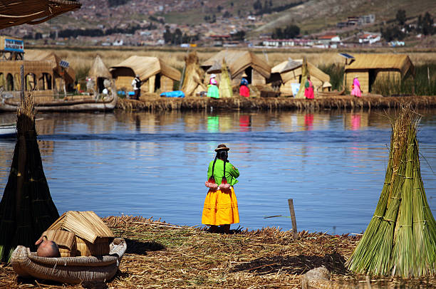 Los Uros Islands, Peru - December 12,2009: Peruvian woman standing  near Lake Titicaca and looking other side of the lake. Houses made of reed and the city of Puno in the hills is in the background. Lake Titicaca is the largest lake in South America and it\\'s partially in Peru and partially in Bolivia.One of the main attractions on the lake are the Uros floating islands made of floating reeds (totora reed).