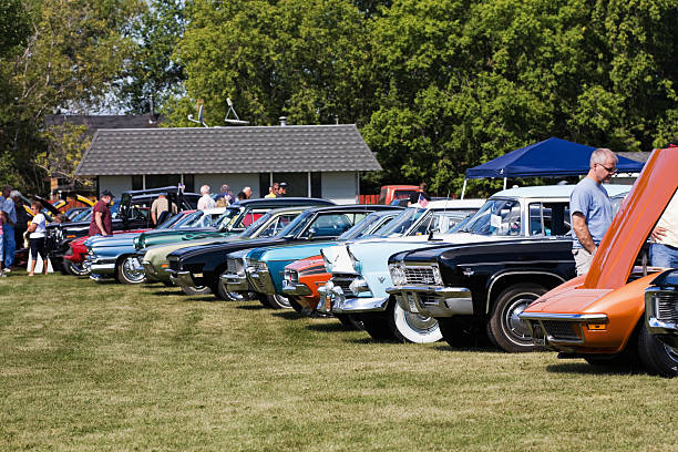 Classic Cars at a Show Fosston, USA - August 27, 2011:  People viewing collector\'s cars that are parked in a row at a classic car show.  In the front, a man is looking under the open hood of an orange Chevrolet Corvette. car show stock pictures, royalty-free photos & images