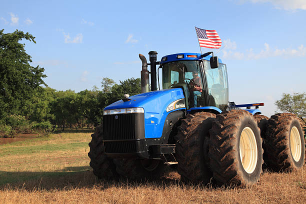 Walters, Oklahoma, USA - June 16, 2010: Patriotic American farmer with a stars and stripes hat driving a New Holland tractor with an American flag flying on top of his cab. Field had muddy places so there\\'s mud on the tires