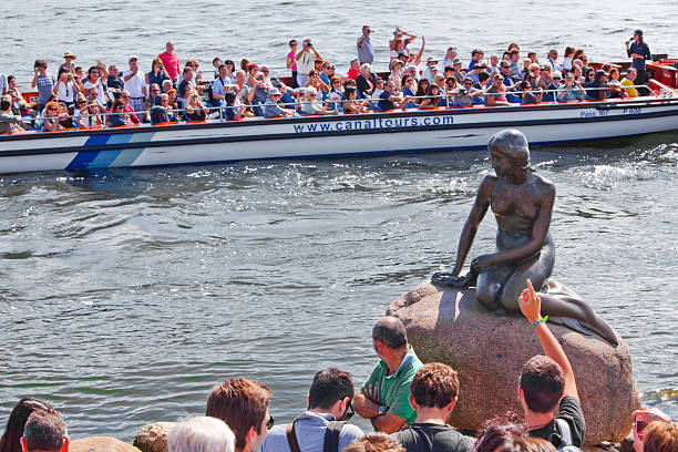 Copenhagen, Denmark - August, 06th 2011: The Little Mermaid statue in Copenhagen. People are standing and looking at the statue. A boat with tourists is passing by.