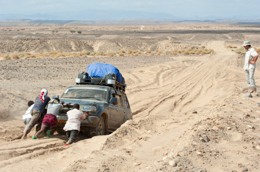 Erta Ale district, Ethiopia - November 27, 2011: A heavy loaded 4WD car of the tourist group got stucked on a sandy road, four Ethiopians are trying to push the car back while a western tourist is watching the scene.The Danakil Desert in the Danakil Depression/Afar triangle in the borderland between Ethiopia, Eritrea and Djibouti is one of the most remote and most extreme regions of the world - it is the lowest point in Africa (- 155 metres/- 550 ft below sea level).