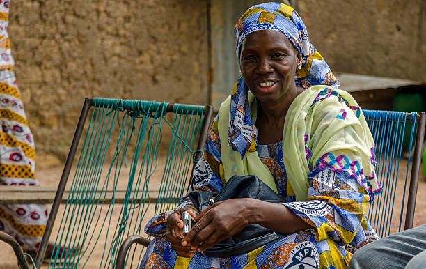 African woman smiling Bamako, Mali – August 18, 2013: An African woman smiling whilst sitting on a chair in front of her house with her typical African clothing and her phone in her hand. Mali stock pictures, royalty-free photos & images