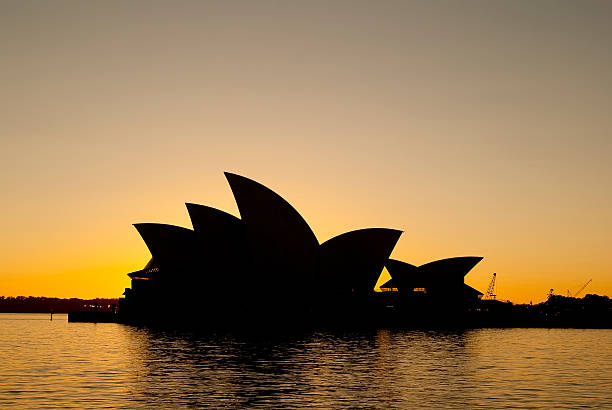 Sydney, Australia - September 14, 2011: The Sydney Opera House, seen from The Rocks, is silhouetted by the rising sun against a clear orange and purple sky.