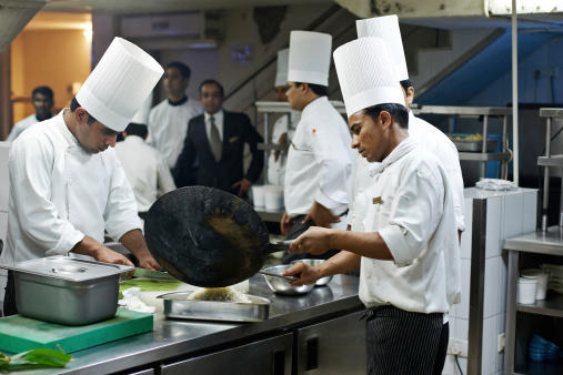 Delhi, India - November 12, 2010: Two Indian hotel chefs preparing food in a busy Delhi hotel kitchen. Four otrher chefs and maitre d'hotel can be seen standing waiting in the background