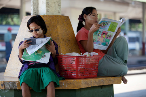 Yangon, Myanmar - February 17, 2011: Young Burmese women sit with a basket full of apples, on a seat curved around a pillar, reading newspapers, waiting for a train on Yangon station. The Yangon central railway station is the largest station in Myanmar, the design based on Burmese style architecture. The trains mostly connect only the major cities and are largely government run. There are only two private companies running trains, the quality of which is better than the government run trains.