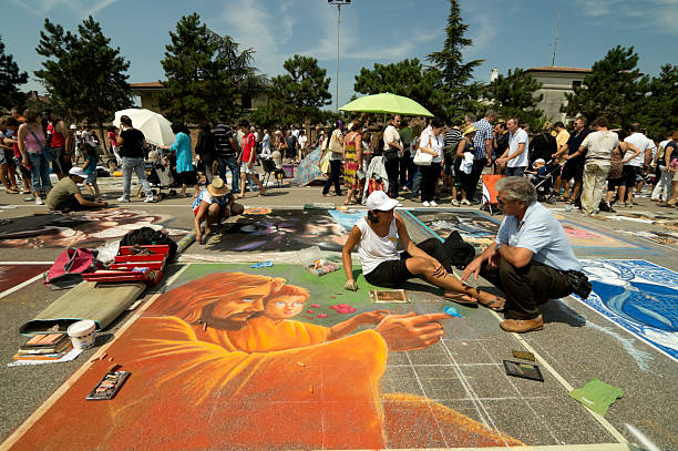 Grazie di Curtatone, Mantova, Italy - August 15, 2011: Madonnari creating their artworks with colored chalks on the churchyard of Santa Maria delle Grazie sanctuary. On the foreground a Jesus with a child is painted with attractive shades of orange. A lot of people admires the street artist and their paintings.Every year on the 15th of August are nearly 200 the challk artists arriving from all over the world to realize their picture meeting together and honoring the Assumption of the Virgin Mary.