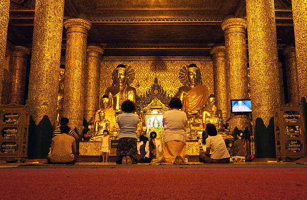 Yangon, Myanmar - March 3, 2011: People kneeling and praying together in an almost completely gold painted prayer room in the famous Shwedagon Pagoda. The famous Shwedagon Pagoda (also known as the Golden Pagoda) is the most sacred pagoda for Buddhists and is said to be 2500 years old. It enshrines relics from the past 4 Buddhas - 8 hairs from Gautama being the most sacred.