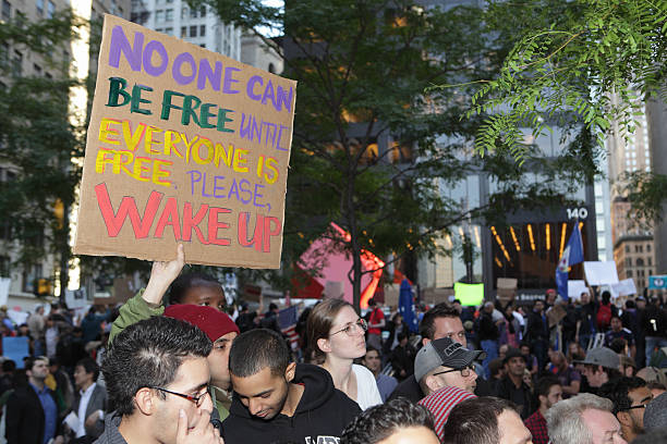 New York, NY, USA - October 7, 2011: An Occupy Wall Street protestor displays his placard in the main camp area while others listen to an address by Jeffrey Sacks of Columbia University's Earth Institute. Zuccotti Park, the location of the camp and rally, is filled with more than 2,000 people taking part in the event and visitors curious to find out more. On September 17, 2011, a group of peaceful protestors set up camp in Zuccotti Park, also called Liberty Park, as part of the Occupy Wall Street campaign to protest corporate influence on democracy, address a growing disparity in wealth, and the absence of legal repercussions behind the recent global financial crisis. Zuccotti Park is in the heart of the Lower Manhattan Financial District, adjacent to Liberty Street, three blocks from Wall Street.