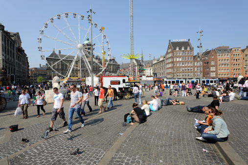 Amsterdam, the Netherlands - April 22, 2011: View at a carnival with a big ferris wheel on Dam Square in Amsterdam. People on the forefront are relaxing in the sun. Behind the ferris wheel is the Royal Palace. Dam Square lies in the historical center of Amsterdam and is one of the city's most well known places. It's a meeting place and a location for many events.
