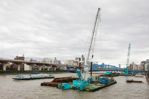 Tokyo, Japan - May 30, 2011: A construction crane mounted on a floating barge works near the seawall of the Sumida River adjacent to the Asakusa area of Tokyo. A Himiko Water Bus passes by in the background. The Sumida River is a river which flows through Tokyo, Japan. It branches from the Arakawa River at Iwabuchi and flows into Tokyo Bay.