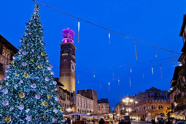 Verona, Italy - December 18, 2009: people in Piazza delle Erbe walking through the market stalls during the Christmas celebrations. Also the medieval Torre dei Lamberti, the tallest building in Verona, is decked out for Christmas.