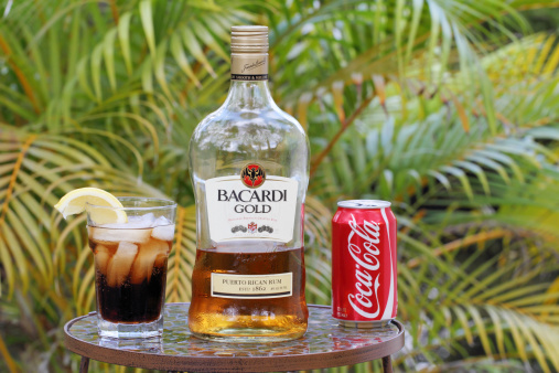 West Palm Beach, USA - March 16, 2011: An outdoor product shot of a half empty Bacardi Gold Rum bottle and an open Coca Cola can shown with a rum and coke cocktail on ice made with those ingredients. Tropical palms are in the background.