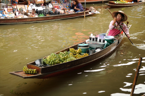 Bangkok, Thailand - November 22, 2008: A woman selling banana and some local drink carefully rows her boat along the floating market in search of customers.  The Damnoen Saduak floating market is the most famous amongst all and is located around a 100kms southwest of Bangkok. The market - beginning at 7.30am and closing by 12.30pm, is one of the most popular places to see in Bangkok. People can buy everything from fresh fruits, vegetables and cooked food to souvenirs. The market is located on a canal dug by King Rama IV to encourage the economic growth of his country. There is a dense population of people living on both sides of the canal, mainly agriculturists by occupation, growing fruits and vegetables of many types which they sell fresh everyday on the floating market. Boats (man powered and motor) are used to navigate through the market and houses of the people living along the canal.