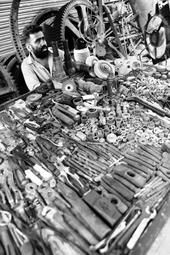 Karachi, Pakistan - June 17, 2010: A middle age Pakistani man sitting by his stall selling used tools and industrial cogwheels outside a closed shop in Shershah Kabari Market (scrapyard) in Karachi, Pakistan. Located in old city area of metropolis Shershah's junkyard is home of few hundreds shops dealing in scrap items ranging from auto parts to industrial junk imported from developed world to recycle in foundries or to give another lease of life through this market.