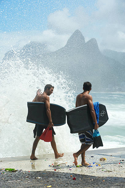 Two Brazilian Surfers Carry Bodyboards in Rio de Janeiro Rio de Janeiro, Brazil - March 16, 2011: Two Brazilian surfers walk on the Ipanema boardwalk at Arpoador, carrying bodyboards and fins next to a crashing wave. During winter swells surfers flock to Arpoador, a famous surf break between Ipanema and Copacabana. two brothers mountain stock pictures, royalty-free photos & images