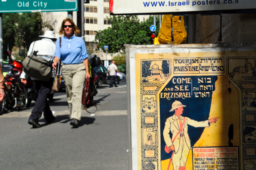 Jerusalem, Israel - October 19, 2010: Pedestrians walk past an antique tourism poster for sale outside a shop on a Jerusalem street. Written in Hebrew and English, it uses  both the terms Palestine and Erez Israel.