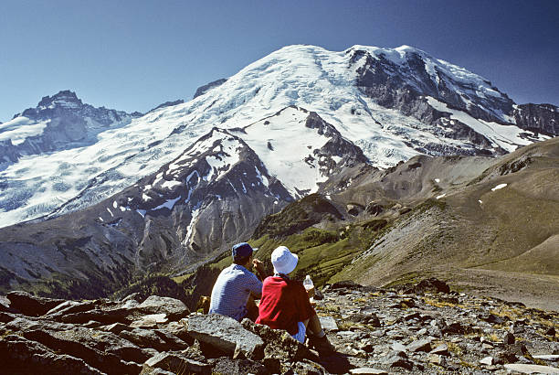Hikers Looking at Mount Rainier Mount Rainier National Park, Washington, USA - August 12, 1995: Two hikers are sitting on the rocks of Burroughs Mountain eating lunch and enjoying the view of Mount Rainier. jeff goulden mount rainier national park stock pictures, royalty-free photos & images