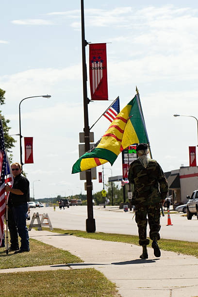 Fosston, USA - August 27, 2011:  A man with long white hair in a ponytail, wearing a beret and camouflage army fatigues, is walking on a sidewalk carrying a Native American Vietnam Veteran flag.  Standing on the grass next to the sidewalk is a man, who is a member of the Patriot Guard Riders, holding an American flag.  He is in a flag line, prior to the funeral of an American soldier who was killed in Afghanistan.  Traffic on US Highway 2, pedestrians, streetlights with signs and banners, and a gas station can be seen in the background.