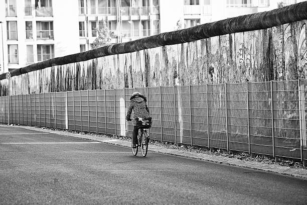 donna in bicicletta lungo le rovine del muro di berlino, germania - east germany berlin germany graffiti wall foto e immagini stock