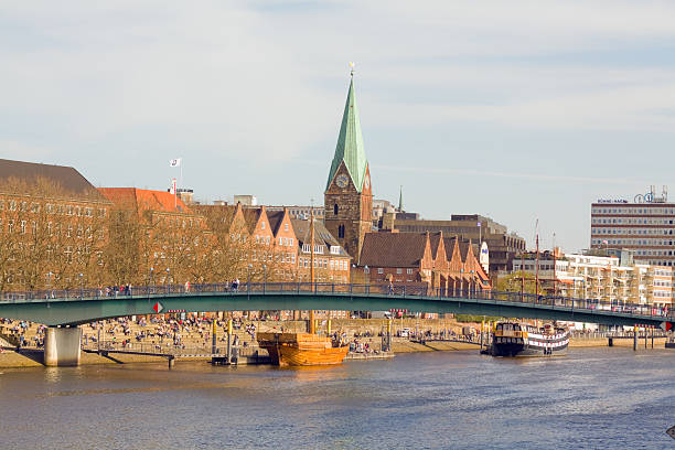 Bremen, Germany - April, 2nd 2011: Promenade Schlachte in Bremen, popular waterfront district at river Weser. A lot of people are walking around, sitting on huge steps at promenade or are passing bridge Teerhof BrAcke. In background is medieval church St. Martini. At promenade are two old sailing ships used as restaurants. At right side is Hilton hotel.