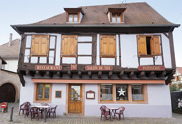 Begheim, France- December 8, 2011: Restaurant in medieval building, in the small village of Begheim, Alsace, France. The house is half-timbered and the rendering is painted lilac. Its gabled roof is actually asymetrical. This is one of the bright colours typical of the local houses. The signs on the restaurant show that it also offers patisserie (cakes and pastries) On the garden wall to the right of the image is the figure of a chef and typical menu signs are in the window and posted on the wall. Berheim is typical of the medieval villages on the wine route through the Rhine Valley, in Alsace, with many medieval buildings preserved offering picturesque tourist attractions.