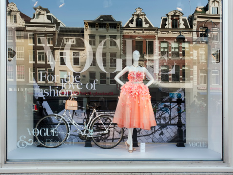 Amsterdam, The Netherlands - March 24, 2012: Vogue Fashion window display in Bijenkorf department store in Amsterdam. Mannequin with dress and bicycle in the window, behind the reflection of old traditional Dutch row houses from across the street.