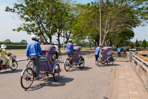 Hue, Vietnam - April 6, 2006: Three Rickshaws drive through the streets of Hue. Each has a passenger on board. Workers can be seen watching the event from the shade in the background.