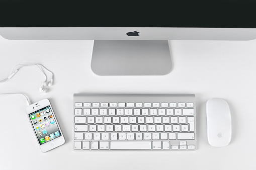 Budapest, Hungary - September 3, 2011: White iPhone 4 with in-ear headphones, Apple keyboard, iMac and Magic Mouse on a white table.