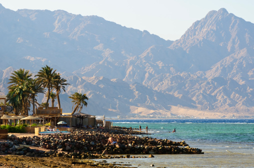 Dahab, Egypt - May 29, 2010: Rugged desert mountains meet the sea at this resort town in the Sinai Peninsula on the Gulf of Aqaba. Swimmers and sunbathers are beside the sea, and the buildings are part of a guesthouse and restaurant.