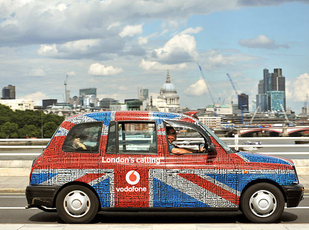 London, UK - July, 22nd 2011:A london Taxi cab with passengers and driver - painted in British Flag design with an advertising slogan for Vodafone, crosses Waterloo Bridge with a view of the City of London