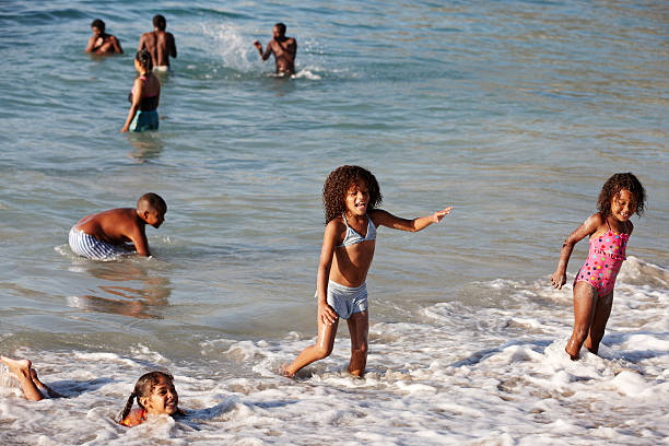 Cape Town, South Africa - January 29, 2012: Children having fun  in the surf on a South African beach. A girl is holding her arms out in a surfing pose, Other children and adults are pictured in the sea in the background. The beach is at Camp's Bay.