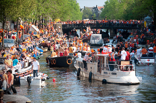 Amsterdam, Netherlands - April 30, 2011: People celebrating Queen's day on and around the canals of Amsterdam. As a tradition, everyone wears orange colored clothing in honor of the royal family, the House of Orange. Party boats sail by as the crowd watch on from the shore.